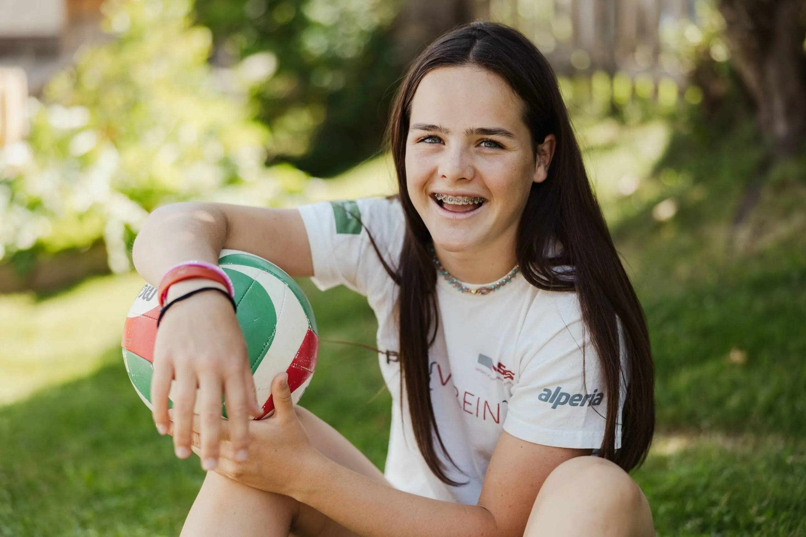 Lena with long dark hair sits on grass outdoors in Alta Badia, Dolomites, holding a multicolor volleyball and smiling.