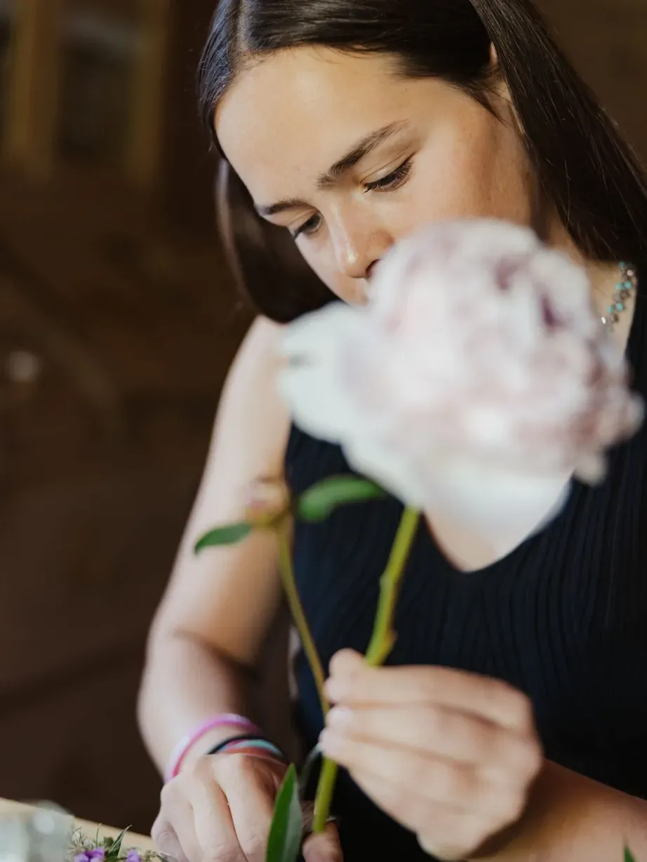Lena with dark hair in a dark sleeveless top looks down at a pale pink flower they hold, with a blurred bouquet in the foreground.