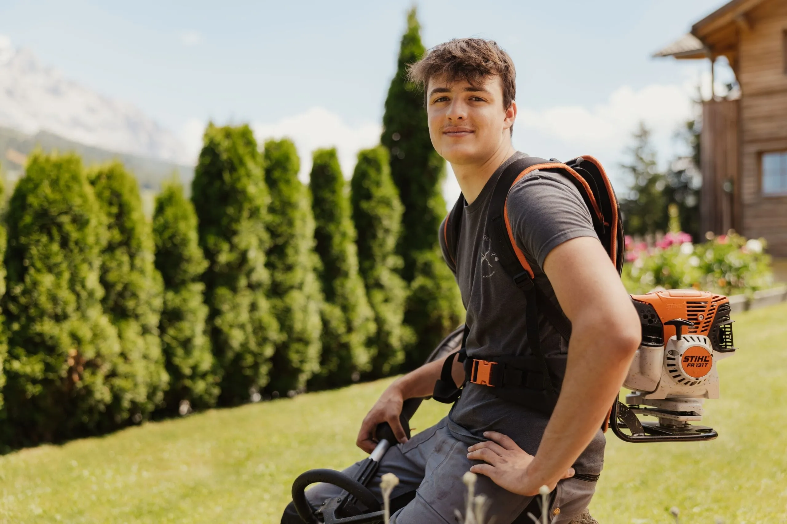 Tommy in a gray tee with an orange backpack leaf blower sits on a lawn, backed by evergreen hedges and a wooden house in Alta Badia, Dolomites.