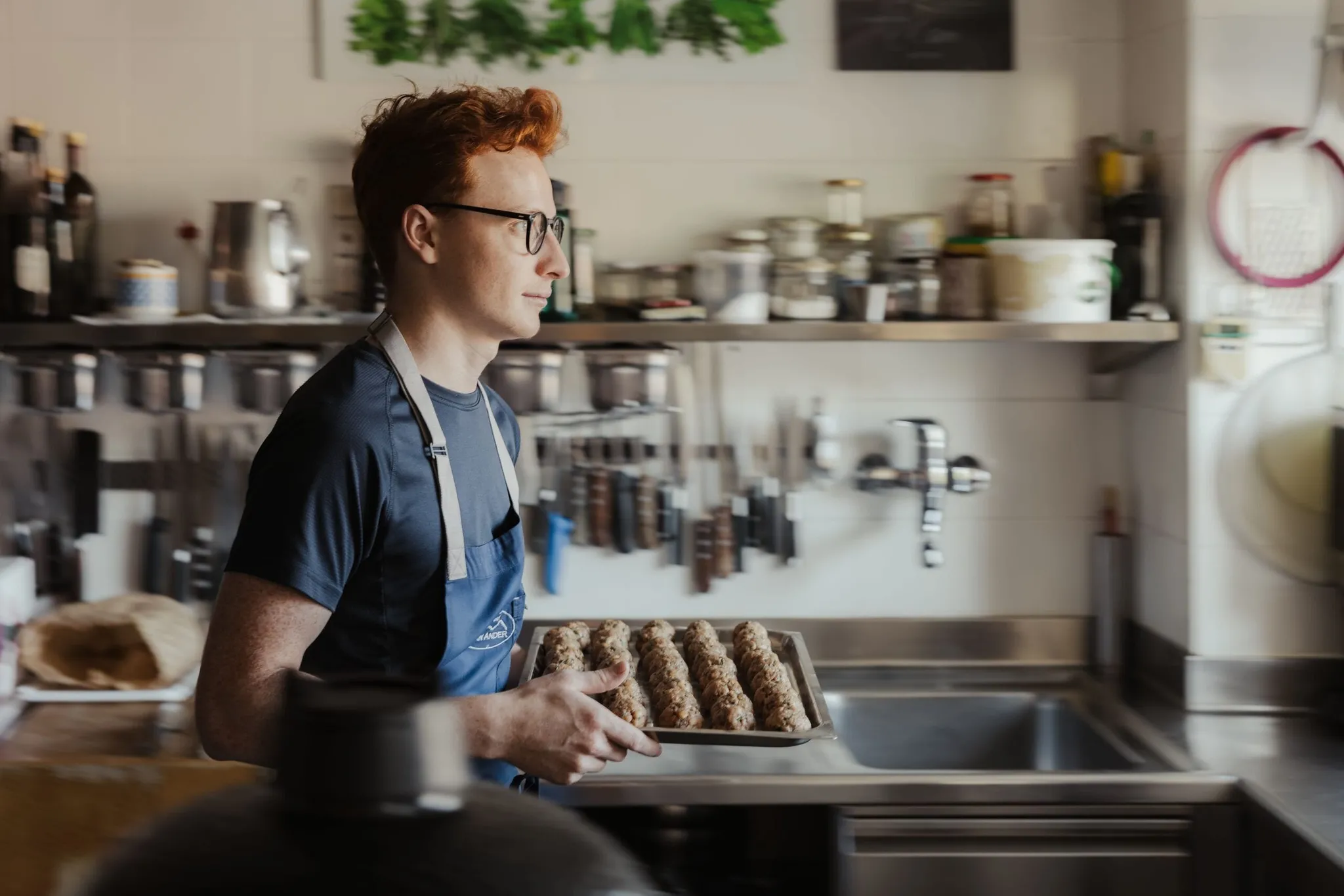Maximilian in a blue apron walks through a kitchen, carrying a tray of sesame-crusted cylindrical snacks.
