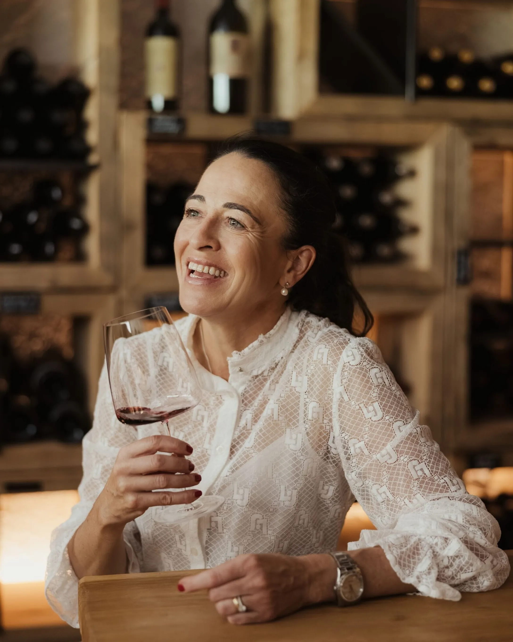 Evelin sorridente, camicetta bianca in pizzo, al bancone con calice di vino rosso; sfondo scaffali di bottiglie in legno.