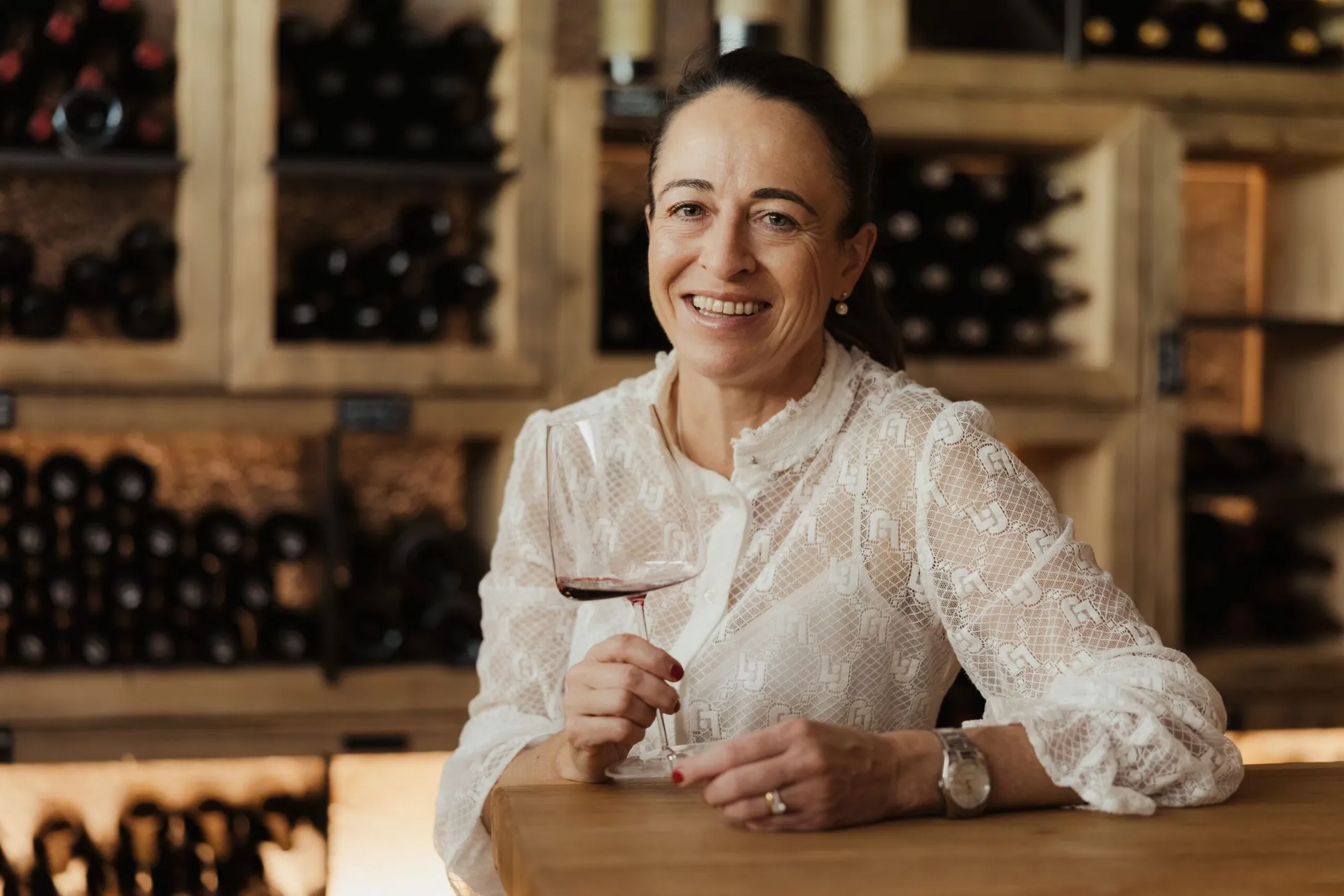 Smiling Evelin in a white lace blouse holds a glass of red wine at a wooden bar, with wine racks in the background.