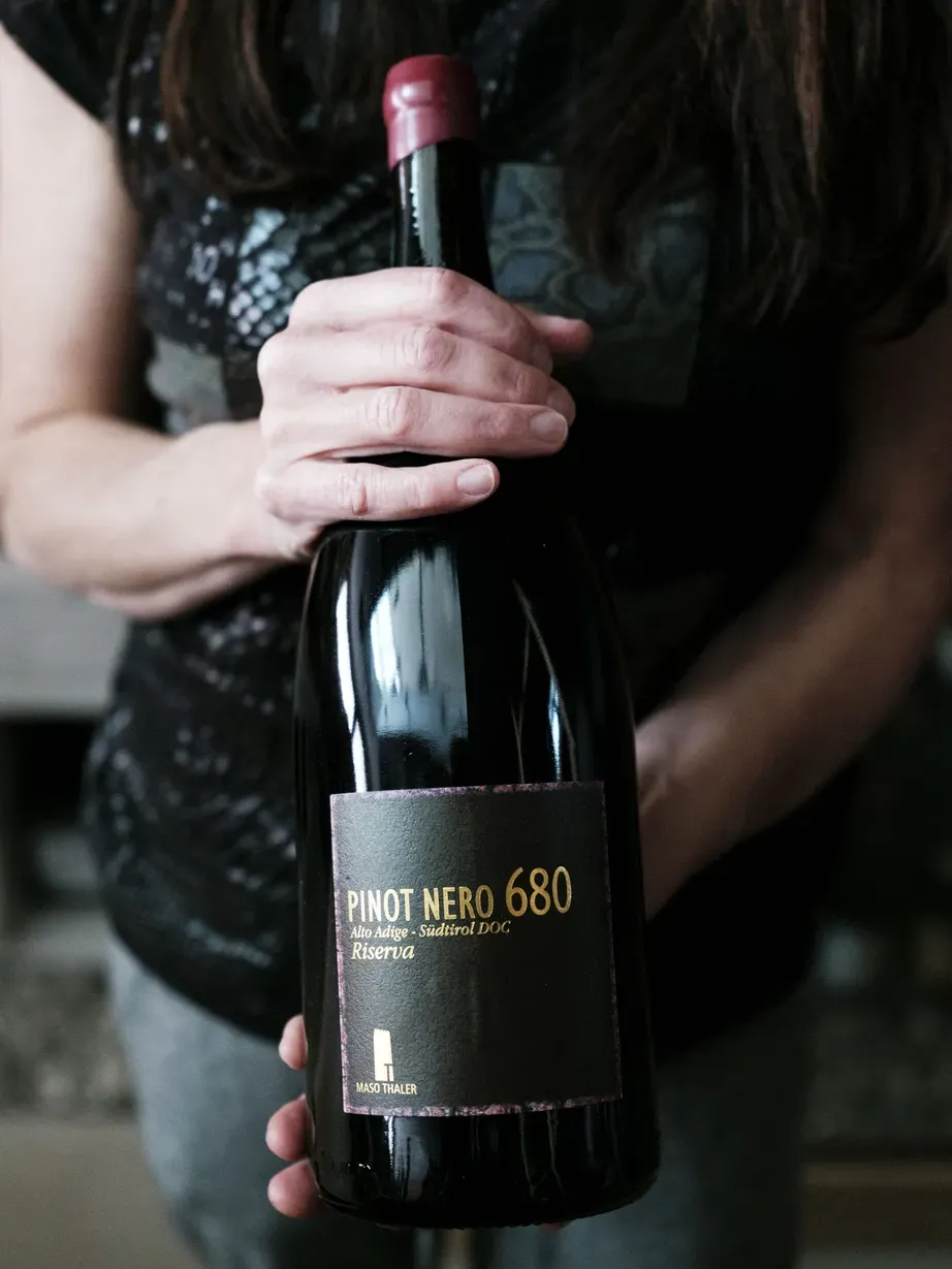 Evelin holds a dark wine bottle indoors; label reads Pinot Nero 680 Riserva with a red foil cap.