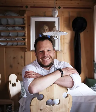 Andrea Irsara sorridente in camicia bianca, appoggiata su una sedia di legno in una stanza rustica dell'Hotel Gran Ander, pareti di legno e tende verdi.