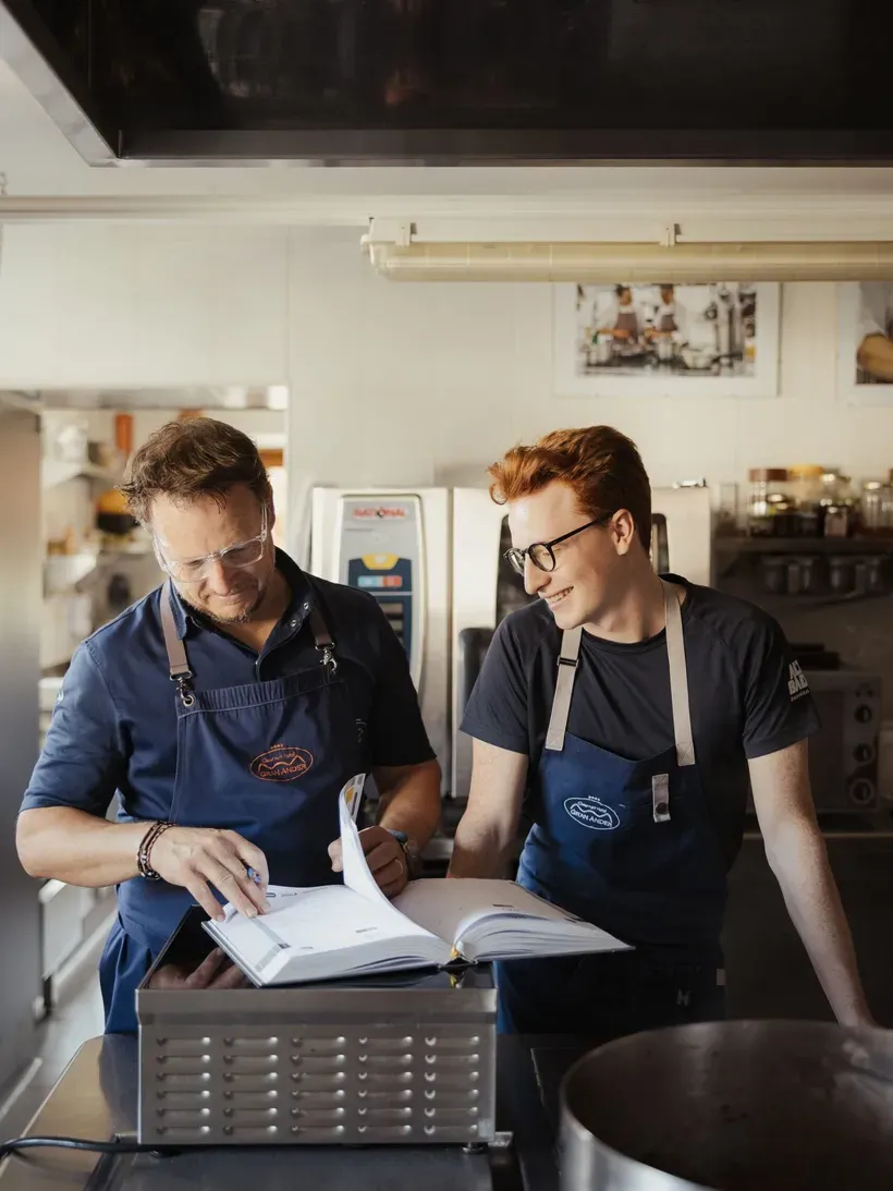 Maximilian e Andrea in blue aprons review an open cookbook in a hotel kitchen, stainless steel counters and equipment around.