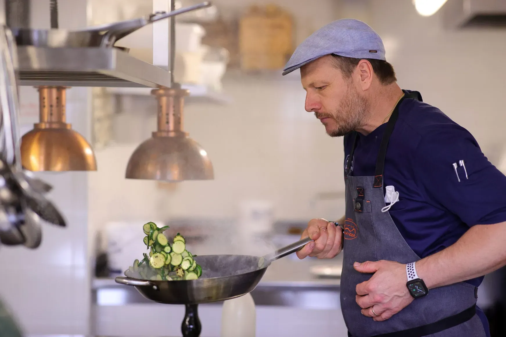 Chef Andrea Irsara wearing a blue shirt and grey apron cooks sliced zucchini in a wok, steam rising as copper heat lamps glow in a bright kitchen.