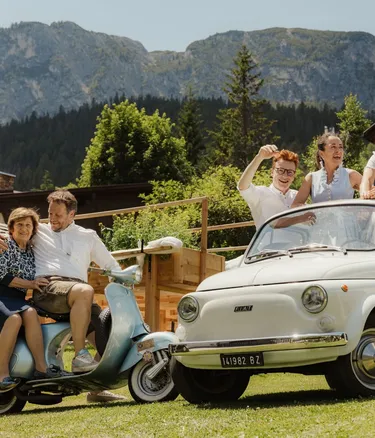 The Irsara family in a vintage Fiat 500 and a blue Vespa, in a sunny garden with a chalet, with the Alta Badia Dolomites in the background.