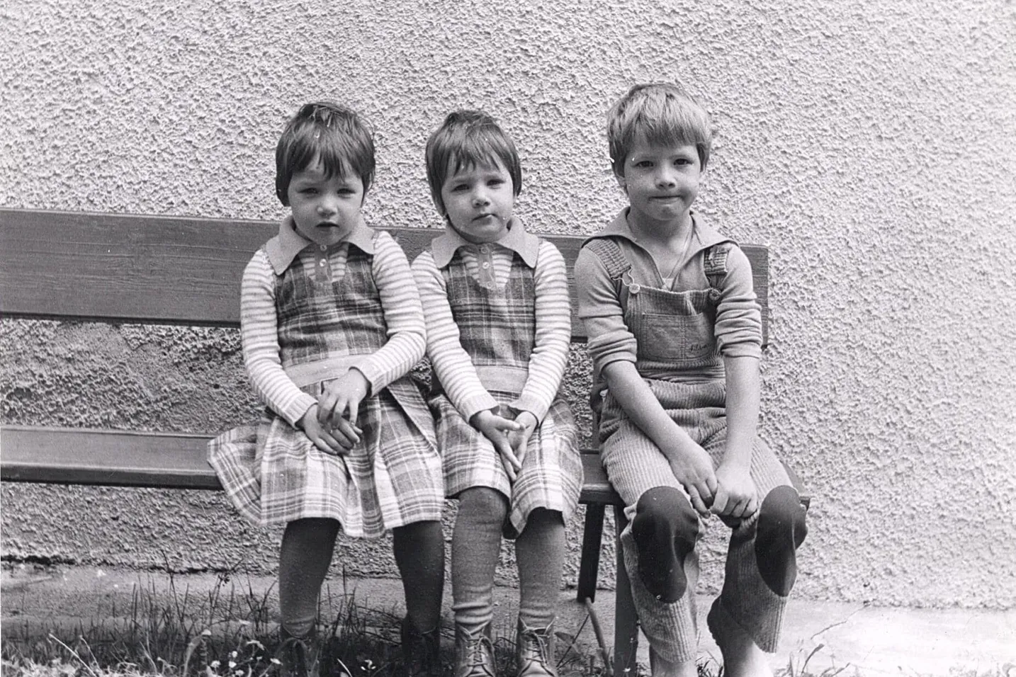 Three children sit on a wooden bench against a textured wall outdoors in Alta Badia, Dolomites; two wear plaid dresses, the third in overalls.