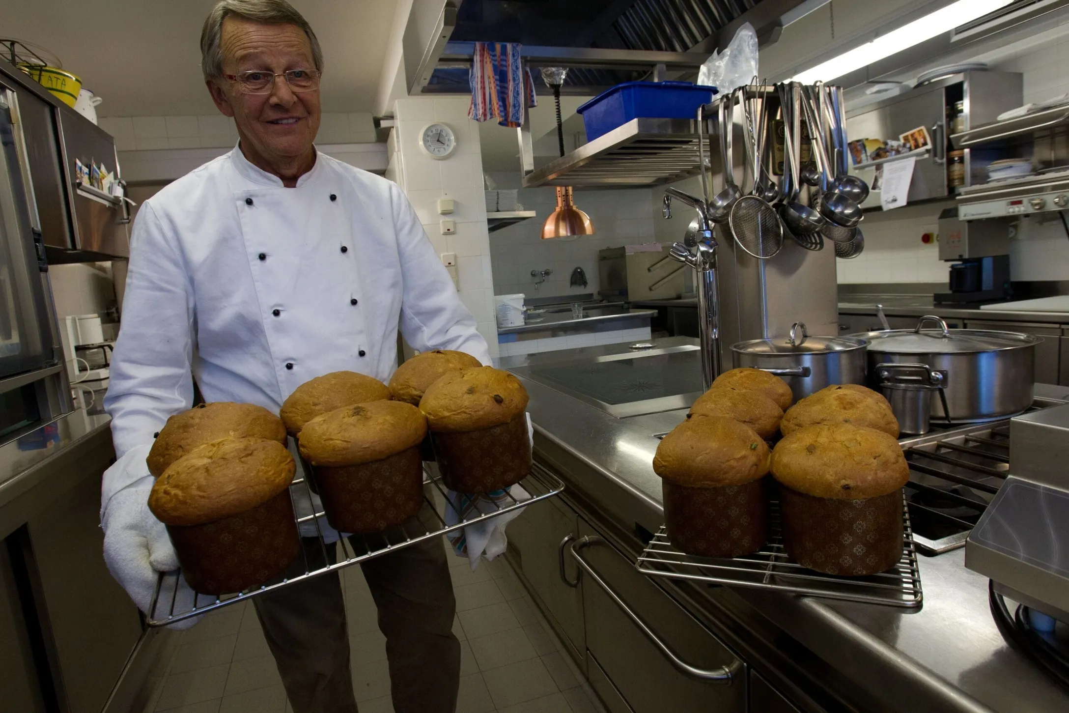 Chef in a white uniform and gloves carries cooling racks of muffins in a stainless steel hotel kitchen.