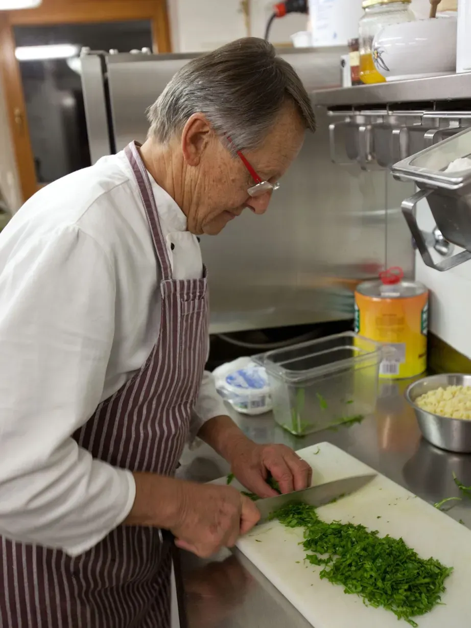 An older person wearing a white chef coat and striped apron chops fresh parsley on a white cutting board in a stainless steel kitchen.