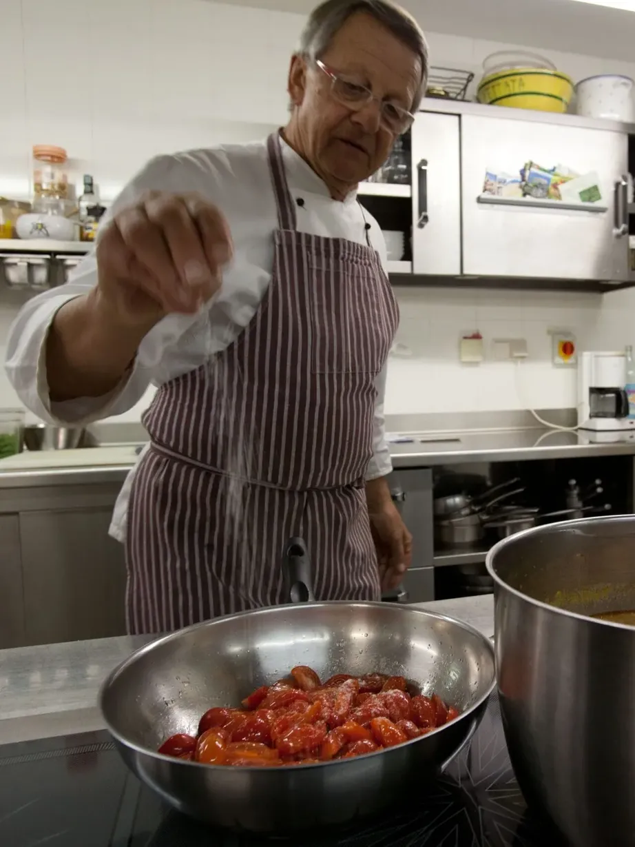 An older chef in a white uniform and striped apron points at the camera, standing in a stainless-steel kitchen with a large pot and a bowl of tomatoes.