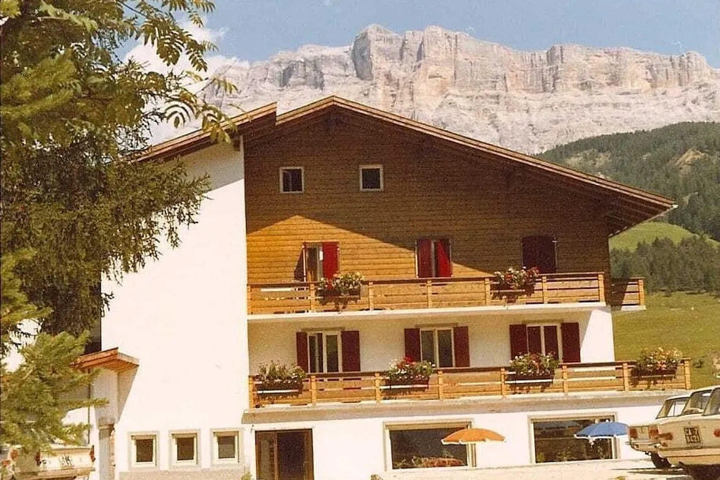 Chalet-style hotel with white base and wooden upper floor, balconies with flower boxes; Alta Badia Dolomites, mountains behind, cars out front.
