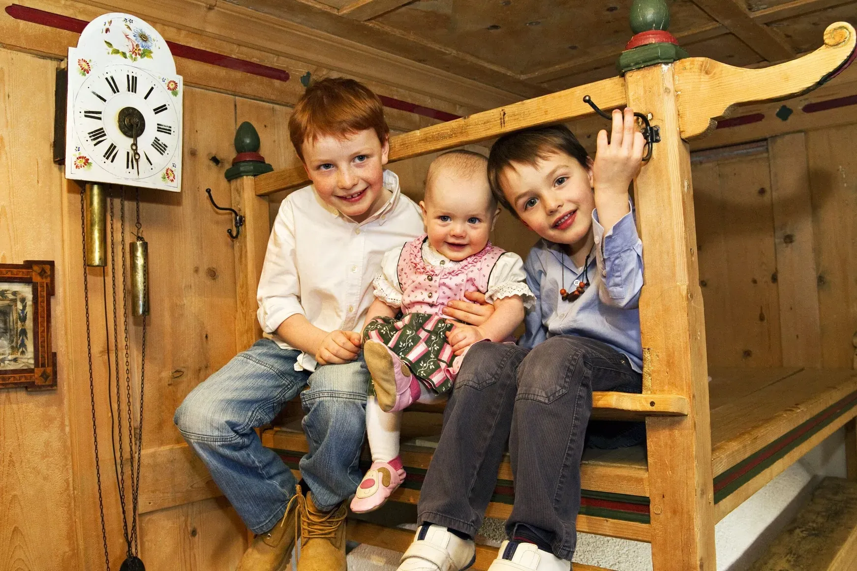 Three children sit on a wooden loft in a rustic room; a vintage floral clock and hanging weights decorate the wall behind them.