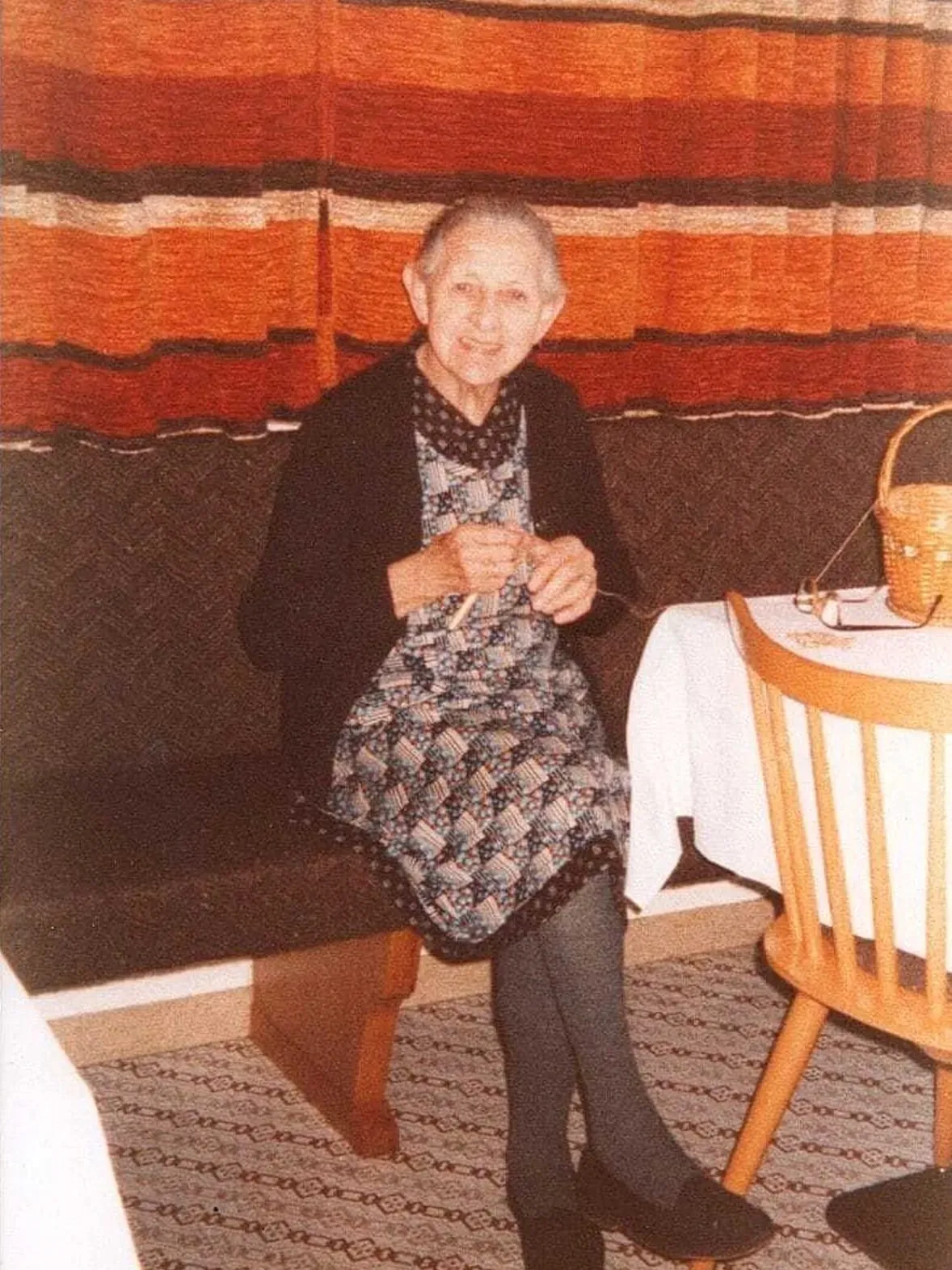 Elderly person seated at a table indoors, knitting, wearing a patterned dress and dark cardigan; orange striped curtain behind.