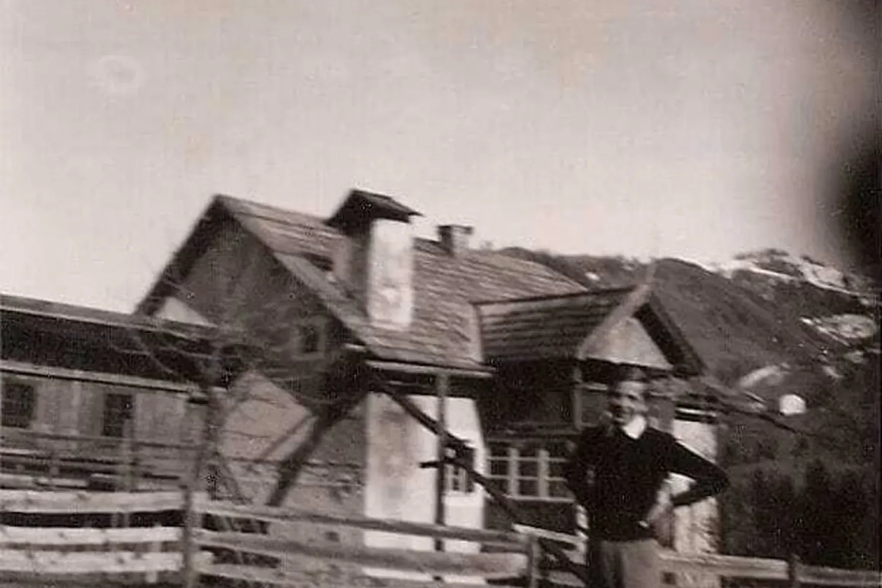 Black-and-white photo of a person standing beside a wooden fence in front of a rustic alpine house with a steep roof, in Alta Badia, Dolomites.